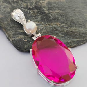 May include: A silver pendant featuring a large, oval-cut, hot pink gemstone. A small, round, white pearl is set above the gemstone. The pendant has a decorative bail for a chain.