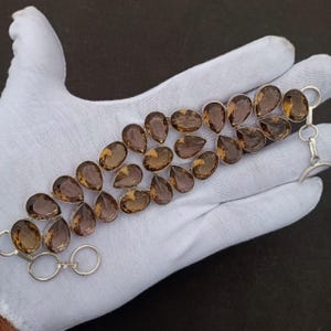 Pode incluir: Uma pulseira de prata com duas fileiras de pedras preciosas de quartzo fumê em forma de gota. As pedras são de cor marrom quente e são engastadas em uma moldura de metal prateado. A pulseira tem um fecho.