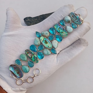 May include: A silver bracelet featuring a variety of blue and green gemstones. The bracelet includes teardrop-shaped, oval, and irregular-shaped stones in shades of turquoise, teal, and iridescent abalone. The bracelet is displayed on a white surface.