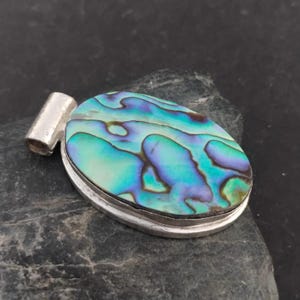 May include: Oval pendant with a vibrant abalone shell inlay, showcasing iridescent blues, greens, and purples. The shell is set in a silver-toned metal frame with a loop for a chain. The pendant is displayed on a dark, textured surface.