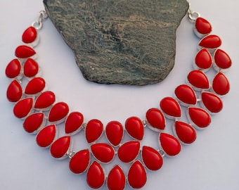 May include: A vibrant red statement necklace featuring teardrop-shaped beads set in a silver-toned metal. The necklace is displayed on a gray stone, showcasing its bold color and design. A striking piece of jewelry.