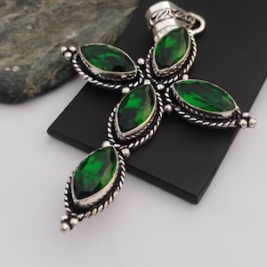 May include: A silver-toned cross pendant featuring five marquise-cut, vibrant green gemstones. The gemstones are set in a detailed, rope-like border. The pendant has a loop at the top for a chain.