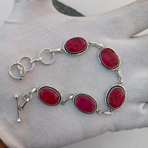 May include: Silver bracelet featuring six oval-cut, vibrant red gemstones. The gemstones are set in individual bezels and connected by silver links. The bracelet has a toggle clasp for secure wear.