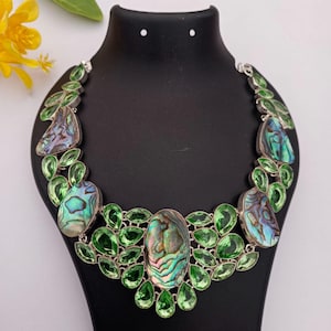 Può includere: Una collana appariscente con pendenti in conchiglia di abalone iridescente e vivaci dettagli di pietre preziose verdi. La collana è incastonata in metallo argentato ed esposta su un manichino nero. Il design incorpora grappoli di pietre verdi a forma di foglia.