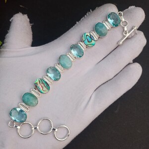 May include: A silver bracelet featuring oval-shaped blue and green gemstones and abalone shell accents. The bracelet has a toggle clasp and is displayed on a white surface. The gemstones are set in a silver link chain.