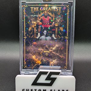 Puede incluir: Una tarjeta coleccionable deportiva dentro de un estuche protector transparente. La tarjeta presenta a tres jugadores de baloncesto, con el texto "THE GREATEST" en la parte superior. El soporte es blanco con el texto "CUSTOM SLABS".