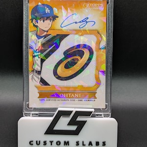 Può includere: Una carta da baseball con l'illustrazione di un giocatore con un berretto blu, con un autografo e una toppa. La carta è racchiusa in un supporto protettivo trasparente, appoggiata su una base bianca con la scritta "CUSTOM SLABS".