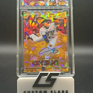 Pode incluir: Um cartão de beisebol de Shohei Ohtani, em uma caixa protetora transparente. O cartão apresenta uma foto de ação do jogador com sua assinatura. O suporte é branco com o texto "CUSTOM SLABS".