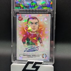Puede incluir: Una tarjeta coleccionable de Cristiano Ronaldo Topps con autógrafo certificado, en un soporte protector. La tarjeta presenta una ilustración de dibujos animados de Ronaldo con la camiseta de Portugal, con su firma. El soporte es blanco con el texto "CUSTOM SLABS".