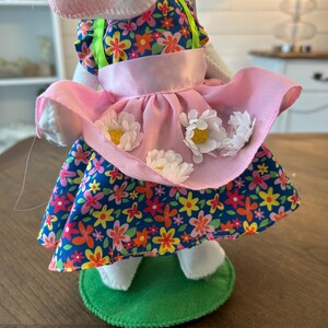 May include: A white bunny doll wearing a floral print dress with a pink apron adorned with white daisy accents. The dress features a blue background with colorful flowers. The doll stands on a green felt base.