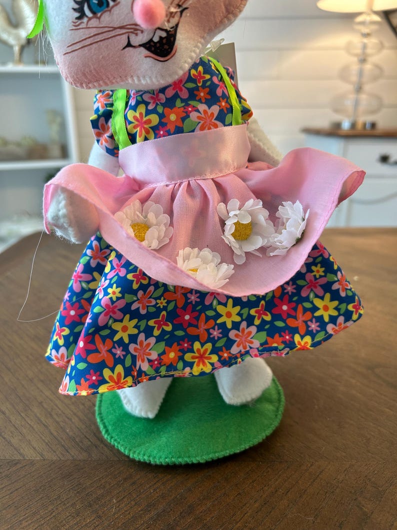 May include: A white bunny doll wearing a floral print dress with a pink apron adorned with white daisy accents. The dress features a blue background with colorful flowers. The doll stands on a green felt base.