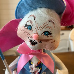 May include: A whimsical Easter bunny doll with a blue hat, pink ears, and a pink bow tie. The bunny wears a blue vest over a floral patterned shirt and holds a small flag. The doll has blue eyes, a pink nose, and white fur.