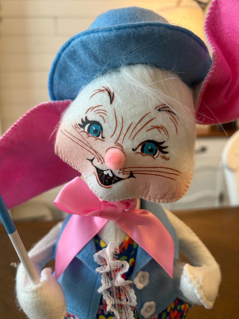 May include: A whimsical Easter bunny doll with a blue hat, pink ears, and a pink bow tie. The bunny wears a blue vest over a floral patterned shirt and holds a small flag. The doll has blue eyes, a pink nose, and white fur.