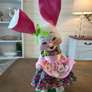 May include: A whimsical Easter bunny figurine with large pink ears and a floral dress. The bunny has a white face with blue eyes and a pink nose. It wears a pink apron with white flower accents and stands on a green base. A green bow adorns its head.