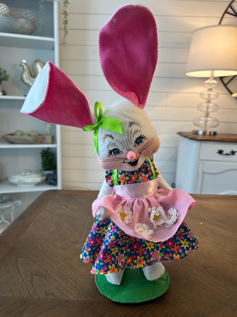 May include: A whimsical Easter bunny figurine with large pink ears and a floral dress. The bunny has a white face with blue eyes and a pink nose. It wears a pink apron with white flower accents and stands on a green base. A green bow adorns its head.