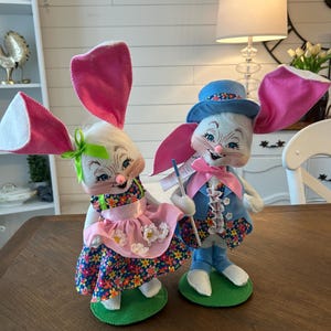May include: Two Easter bunny figurines. One bunny wears a pink floral dress and a pink apron. The other bunny wears a blue suit and a blue hat. Both bunnies have large pink ears and are standing on green bases.