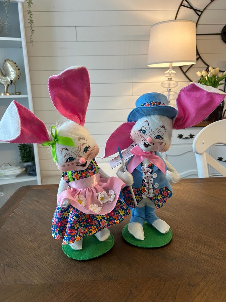 May include: Two Easter bunny figurines. One bunny wears a pink floral dress and a pink apron. The other bunny wears a blue suit and a blue hat. Both bunnies have large pink ears and are standing on green bases.