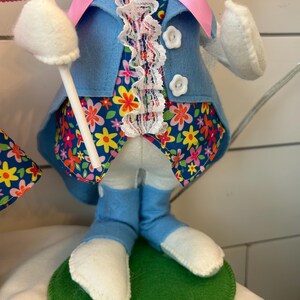May include: A decorative Easter bunny figure. The bunny wears a light blue felt jacket with white buttons, a floral patterned vest, and blue felt pants. The bunny's feet are white, and it stands on a green felt base. A pink bow and white lace trim the bunny's outfit.