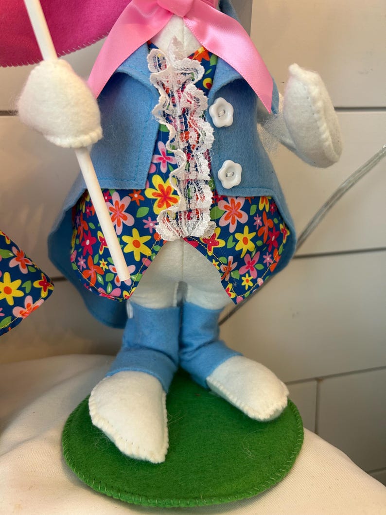 May include: A decorative Easter bunny figure. The bunny wears a light blue felt jacket with white buttons, a floral patterned vest, and blue felt pants. The bunny's feet are white, and it stands on a green felt base. A pink bow and white lace trim the bunny's outfit.