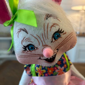 May include: A plush white bunny with pink ears, blue eyes, and a pink nose. The bunny wears a blue floral top and a pink skirt. A green bow adorns its head, creating a whimsical and cheerful Easter or spring-themed decoration.