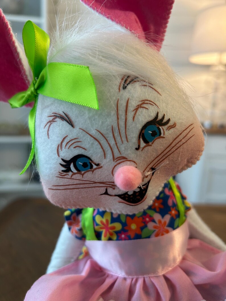 May include: A plush white bunny with pink ears, blue eyes, and a pink nose. The bunny wears a blue floral top and a pink skirt. A green bow adorns its head, creating a whimsical and cheerful Easter or spring-themed decoration.