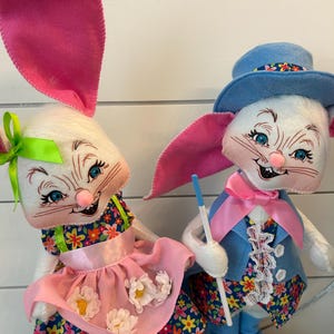 May include: Two plush Easter bunny dolls. One bunny wears a floral dress with a pink apron and a green bow. The other bunny wears a blue suit, a pink bow tie, and a blue hat. Both bunnies have pink ears and are smiling.