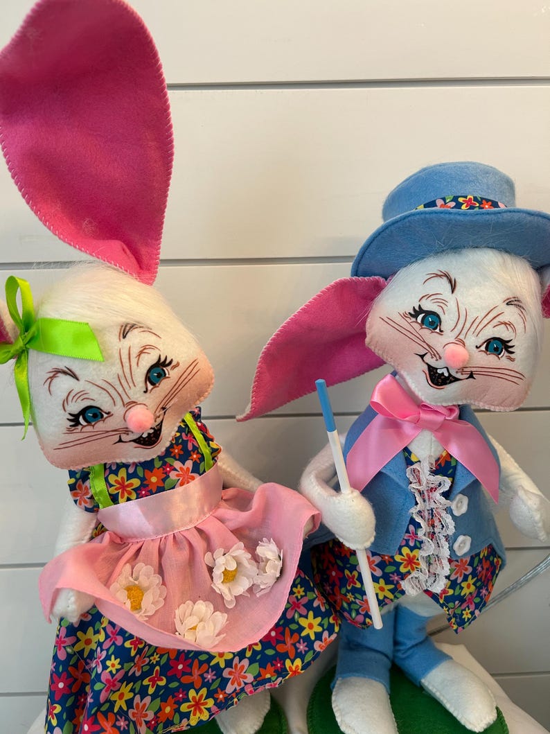 May include: Two plush Easter bunny dolls. One bunny wears a floral dress with a pink apron and a green bow. The other bunny wears a blue suit, a pink bow tie, and a blue hat. Both bunnies have pink ears and are smiling.