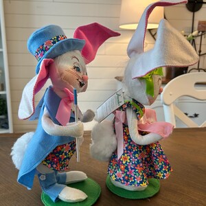 May include: Two Easter bunny figurines. One bunny wears a blue top hat with floral trim, a blue vest, and a floral patterned shirt. The other bunny wears a floral dress and a pink bow. Both have long ears and are standing on green bases.