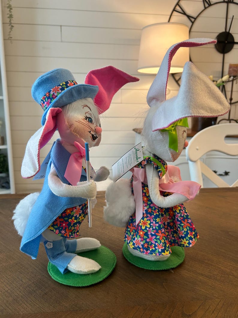 May include: Two Easter bunny figurines. One bunny wears a blue top hat with floral trim, a blue vest, and a floral patterned shirt. The other bunny wears a floral dress and a pink bow. Both have long ears and are standing on green bases.