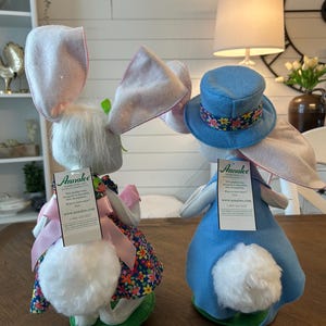 May include: Two Easter bunny dolls. One bunny wears a floral dress and pink bow, the other a blue top hat with a floral band. Both have large pink-lined ears and fluffy white tails. The dolls are on a wooden table.