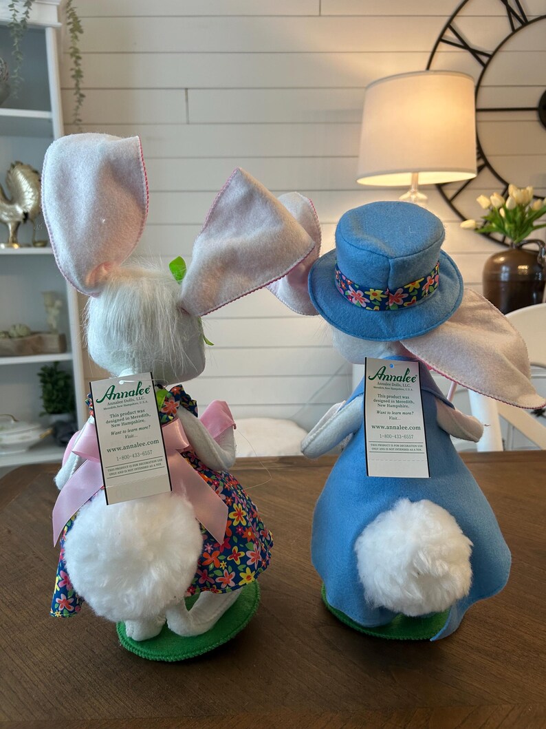 May include: Two Easter bunny dolls. One bunny wears a floral dress and pink bow, the other a blue top hat with a floral band. Both have large pink-lined ears and fluffy white tails. The dolls are on a wooden table.