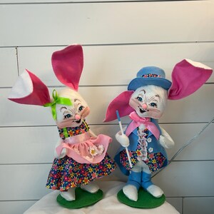 May include: Two Easter bunny dolls. One doll wears a floral dress with a pink apron and a green bow. The other doll wears a blue suit with a matching hat and holds a blue stick. Both dolls have large pink and white ears.
