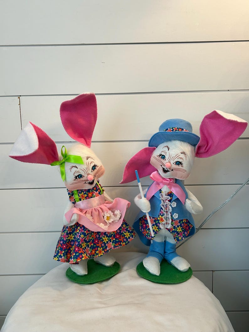May include: Two Easter bunny dolls. One doll wears a floral dress with a pink apron and a green bow. The other doll wears a blue suit with a matching hat and holds a blue stick. Both dolls have large pink and white ears.