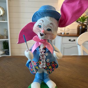 May include: A whimsical Easter bunny figurine. The bunny wears a blue hat, jacket, and pants, with a floral print vest and a pink bow tie. It has large pink ears and holds a blue-tipped wand. The figurine stands on a green base.