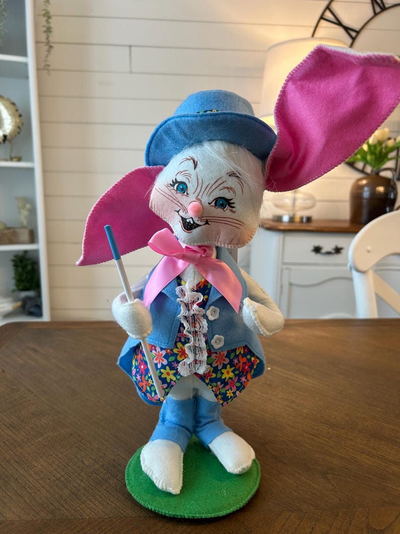 May include: A whimsical Easter bunny figurine. The bunny wears a blue hat, jacket, and pants, with a floral print vest and a pink bow tie. It has large pink ears and holds a blue-tipped wand. The figurine stands on a green base.