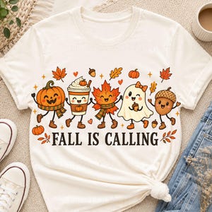 May include: A cream-colored short-sleeve t-shirt featuring a colorful graphic of a smiling pumpkin, a coffee cup, a maple leaf, a ghost, and an acorn. The text "FALL IS CALLING" is printed in black below the characters, surrounded by small stars.