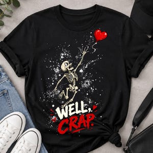 May include: A black short-sleeve t-shirt featuring a graphic of a skeleton reaching for a red heart-shaped balloon. Below the skeleton, the text reads WELL, CRAP in white and red lettering, surrounded by white paint splatter effects.