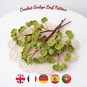 Può includere: Un modello di foglia di ginkgo all'uncinetto con diverse foglie verdi a ventaglio e piccole bacche verdi rotonde e testurizzate attaccate a sottili steli marroni simili a rami. Il testo "Crochet Ginkgo Leaf Pattern" è visibile in alto.