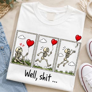 May include: A white t-shirt featuring a three-panel comic graphic of a skeleton. The first panel shows a skeleton sitting with a red heart balloon, the second shows it running, and the third shows the balloon floating away. The text reads Well, shit...