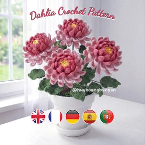 May include: A white ceramic pot holds a bouquet of crocheted dahlias. The flowers are various shades of pink with yellow centres and green leaves. The text "Dahlia Crochet Pattern" is at the top.