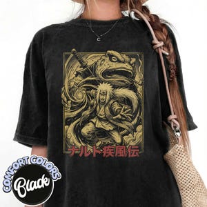 May include: Black Comfort Colors t-shirt with a golden illustration of a character with white hair and a frog. The character is holding a sword and is surrounded by swirling energy. Japanese text is below the illustration.