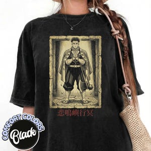 Puede incluir: Camiseta negra Comfort Colors con una ilustración detallada de un personaje en un marco de piedra. El personaje lleva una capa y junta las manos. Texto japonés debajo de la ilustración. La camiseta tiene una etiqueta Comfort Colors Black.