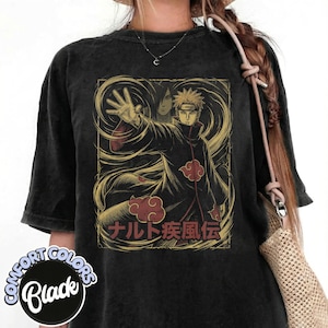 May include: Black Comfort Colors t-shirt featuring a graphic of an anime character in a black cloak with red cloud accents. The design includes Japanese text and a gold-colored background. The shirt has a vintage, worn look.