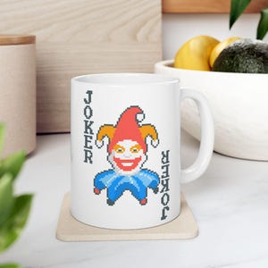 May include: White ceramic mug featuring a pixelated joker design. The joker has a red hat, blue collar, and a smiling face. The word "JOKER" is printed vertically on both sides of the mug. The mug is on a beige coaster.