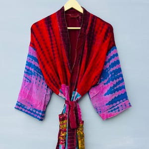 May include: A long-sleeved robe featuring a vibrant tie-dye pattern in shades of red, pink, blue, and purple. The garment includes a matching fabric tie belt at the waist, wide sleeves, and a deep red collar, all displayed on a wooden hanger.