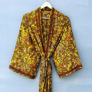 May include: A long-sleeved yellow kimono robe featuring an intricate floral pattern in shades of orange, brown, and green. The garment includes a matching fabric tie belt at the waist and a contrasting dark red border along the front opening and cuffs.
