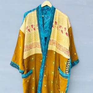 May include: A long-sleeved kimono robe featuring a mustard yellow base with a repeating sun pattern, accented by a bright turquoise blue trim and collar. The upper section displays a cream-colored panel with red leaf motifs and two front pockets.