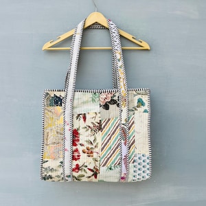 May include: A rectangular quilted tote bag featuring a patchwork design of various floral and striped fabric patterns in shades of red, yellow, green, and blue. The bag has two long shoulder straps with a black and white striped trim.
