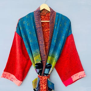 May include: A long-sleeved kimono robe featuring a vibrant patchwork design with contrasting panels of teal, bright red, and orange. The garment includes a patterned collar, wide sleeves, and a matching fabric tie belt at the waist.