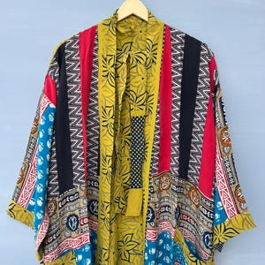 May include: A long-sleeved kimono robe featuring a patchwork design with various patterns and colors including mustard yellow, red, black, and blue. The garment has a wide collar, an open front, and a tie belt, all displayed on a wooden hanger.